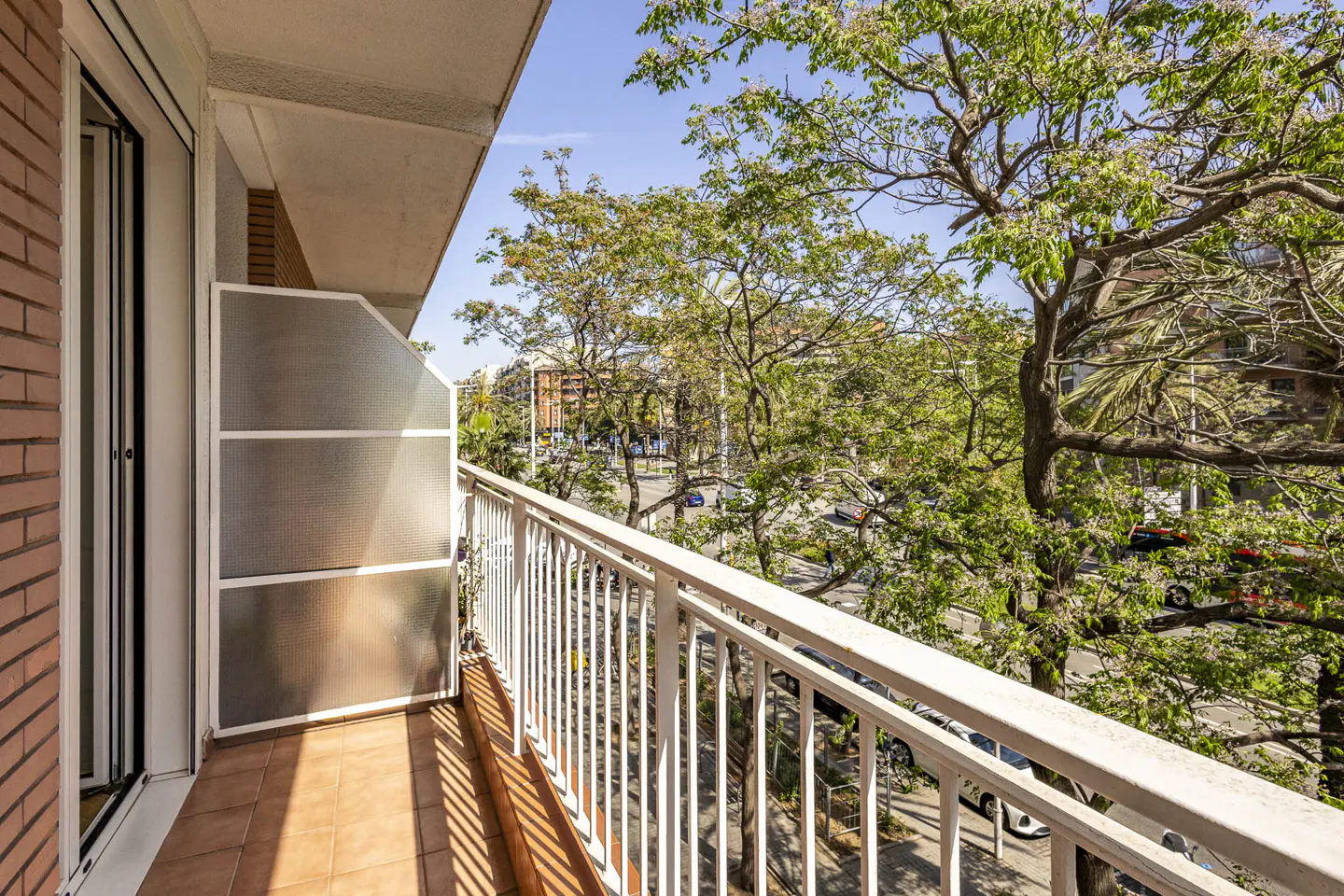 Balcony view with white railings, brown tile floor, and a partial privacy screen. Trees and a street scene are visible in the background.