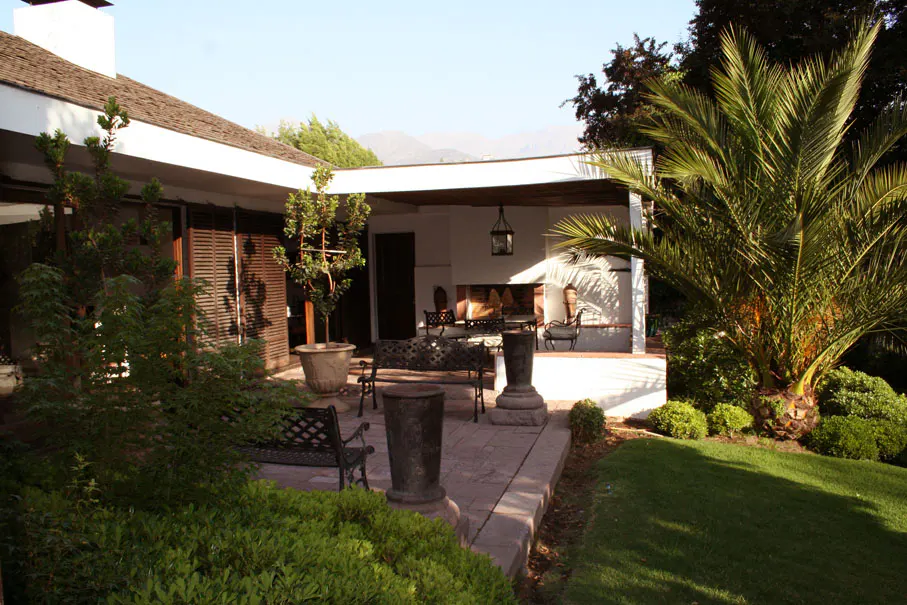 Outdoor patio with a fireplace, bench, and palm tree. The patio is made of stone and has a white roof. Mountains are visible in the background.