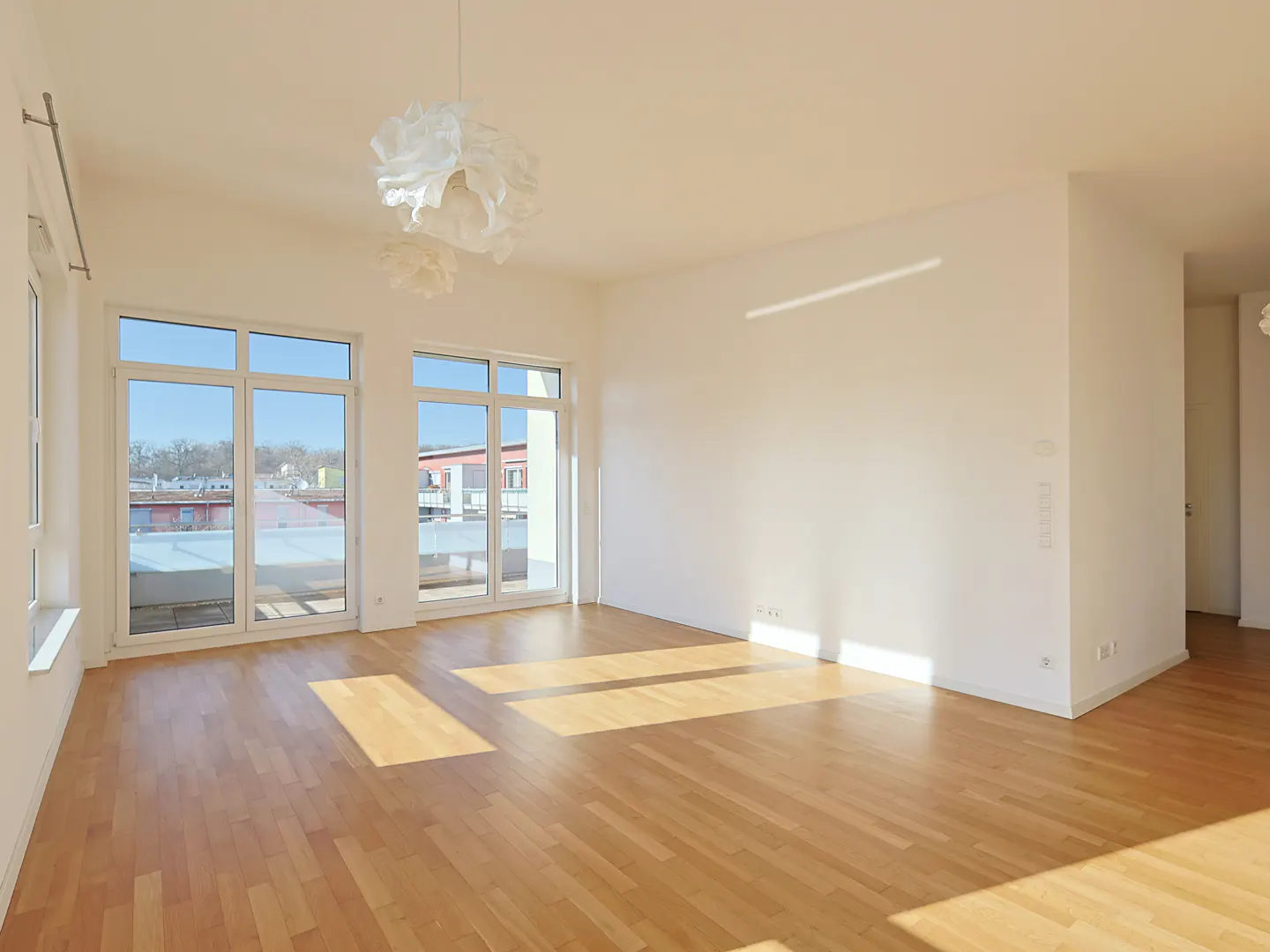 Bright, empty room with light wood floors, white walls, and large windows. Modern white pendant lights hang from the ceiling.