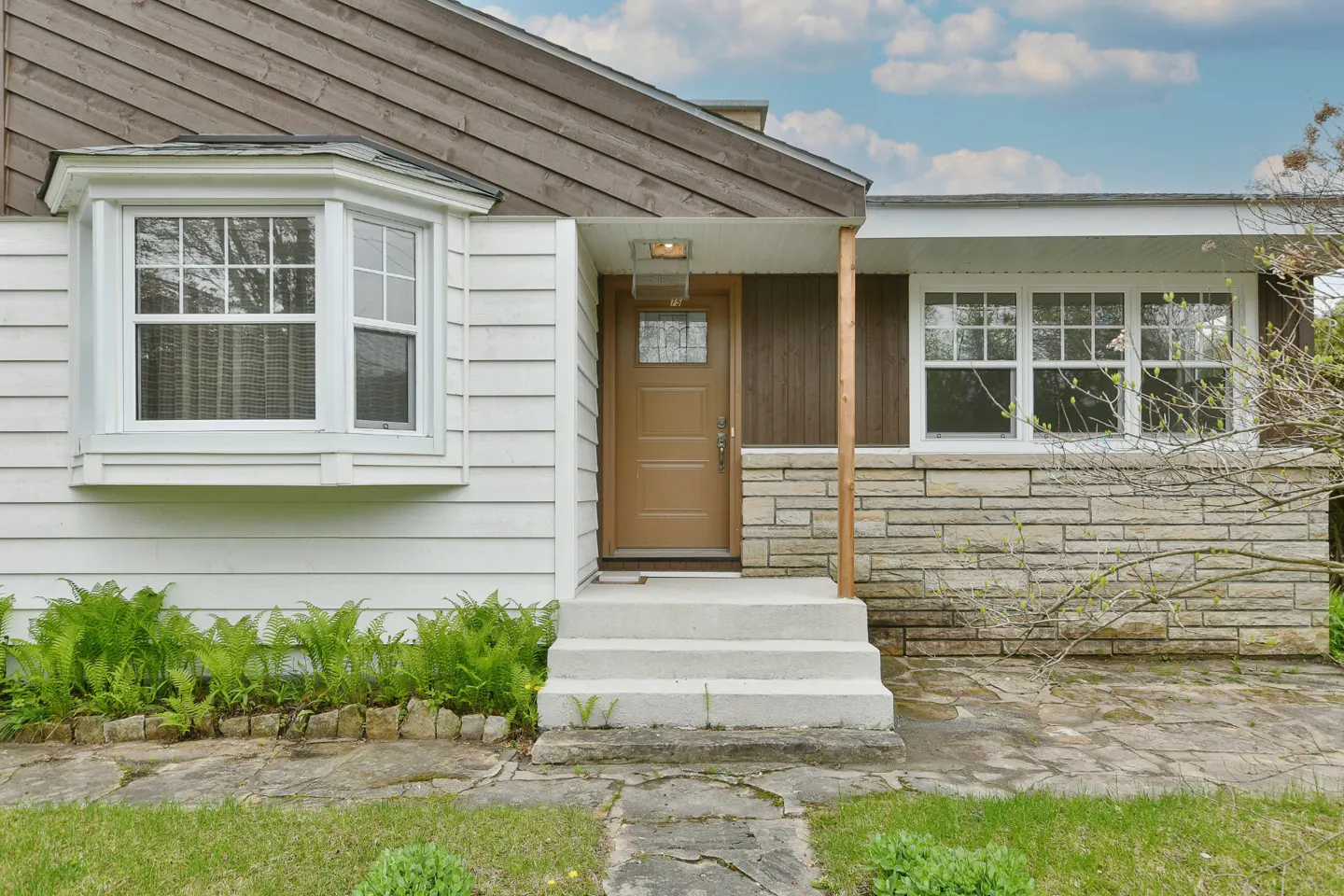 Exterior view of a house with white siding, brown trim, and a stone foundation. A brown front door is centered with concrete steps leading up to it. Ferns grow along the base of the house.