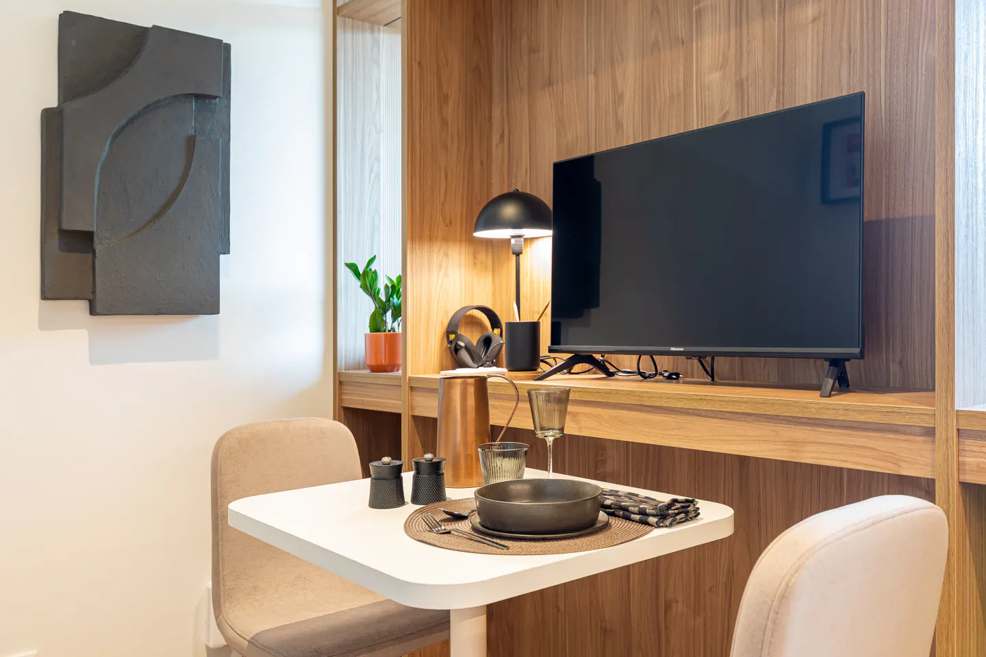 Interior view of a modern dining area with a white table set for one, chairs, and a TV on a wooden shelf. A black abstract art piece hangs on the wall.