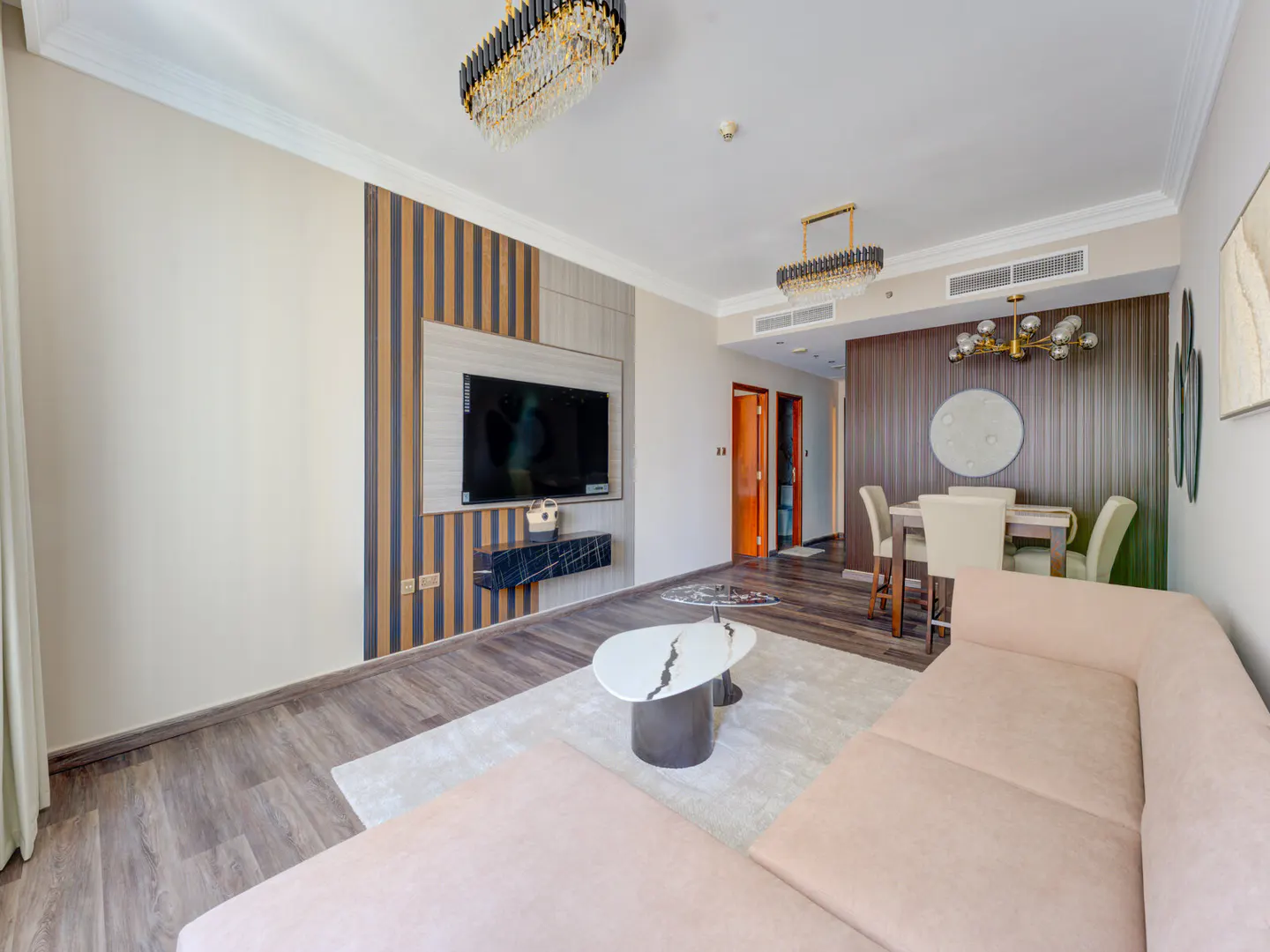 A modern living room with a beige sectional sofa, a marble coffee table, and a flat-screen TV mounted on a striped wall. A dining area is visible in the background.