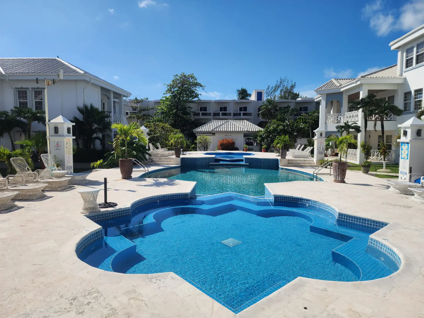 A bright blue geometric pool is surrounded by white buildings and lounge chairs under a clear blue sky.