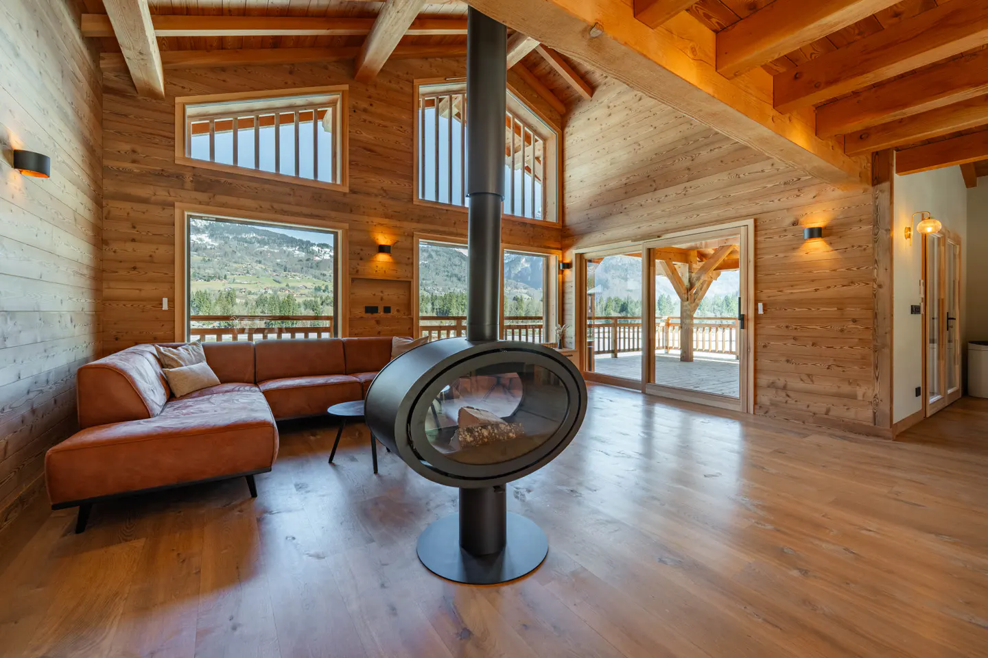 Rustic living room with wood walls, floors, and ceiling beams. A modern black fireplace hangs from the ceiling. Large windows overlook a mountain view.
