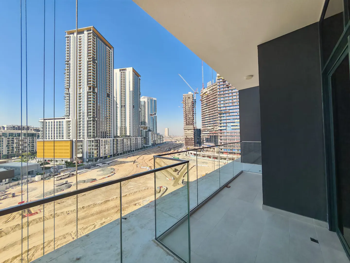 View from a balcony with a glass railing overlooking a construction site and city skyline under a blue sky.