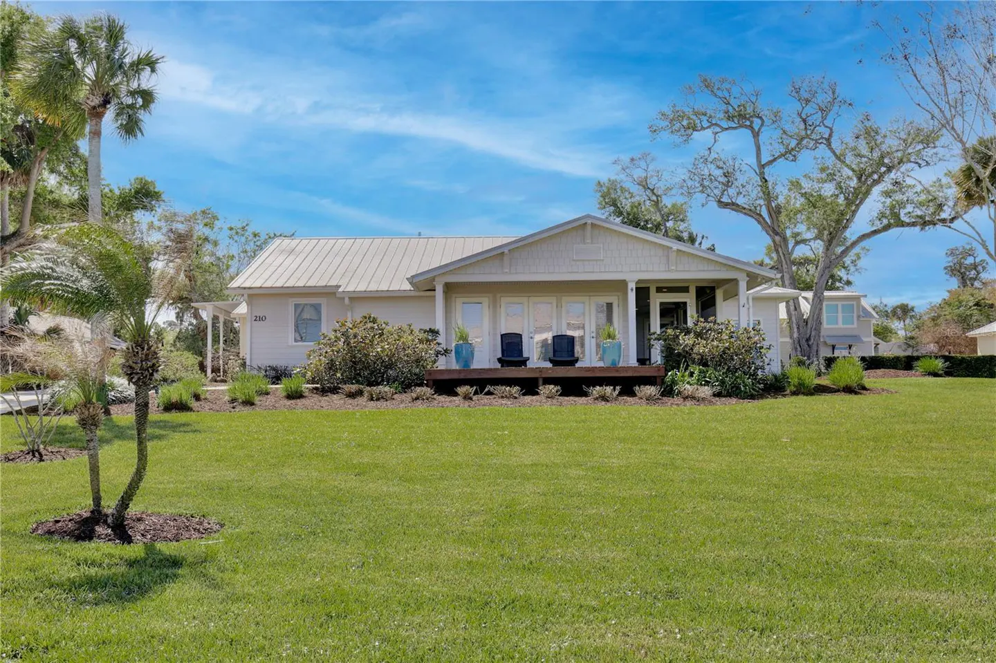 A single-story, light-beige house with a metal roof, a wooden porch with two chairs, and a green lawn.