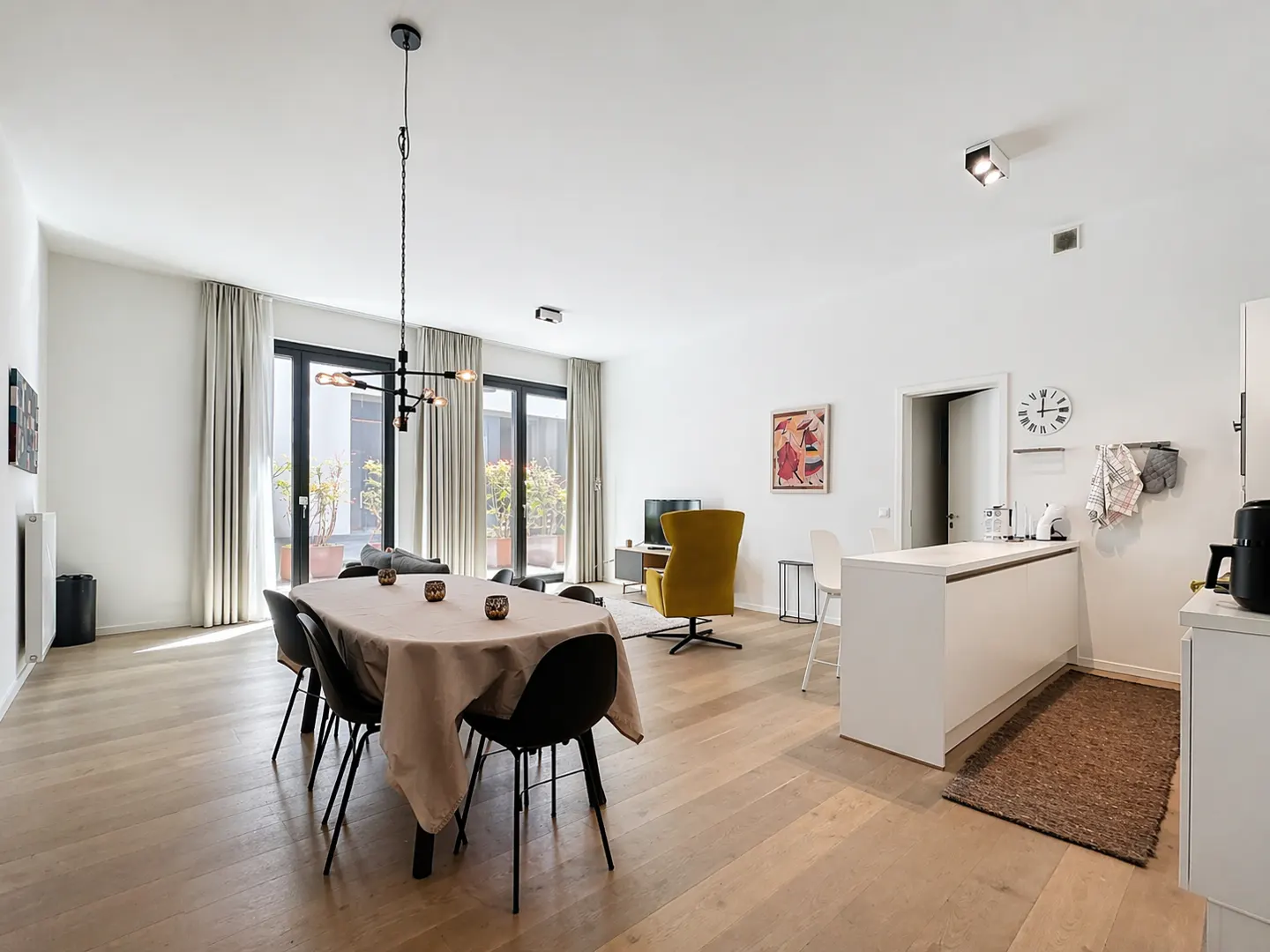 Bright, open-plan living space with wood floors, dining table, kitchen island, and yellow chair. Large windows with curtains.
