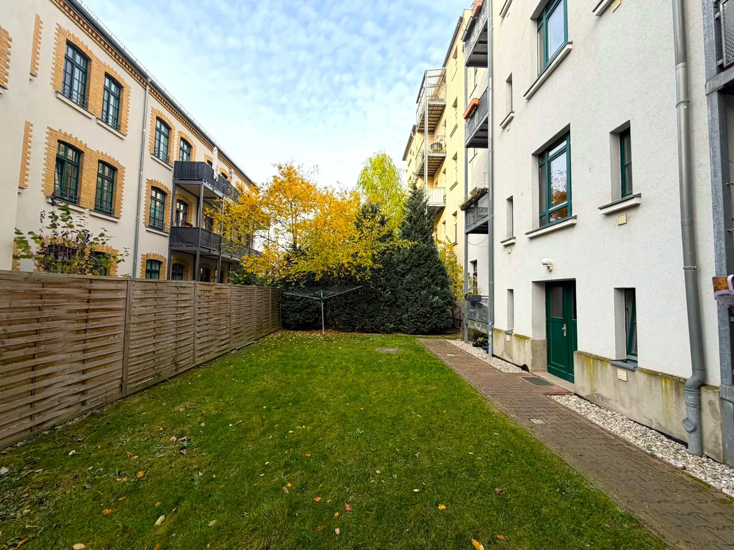 A green lawn is surrounded by buildings and a wooden fence under a cloudy sky.