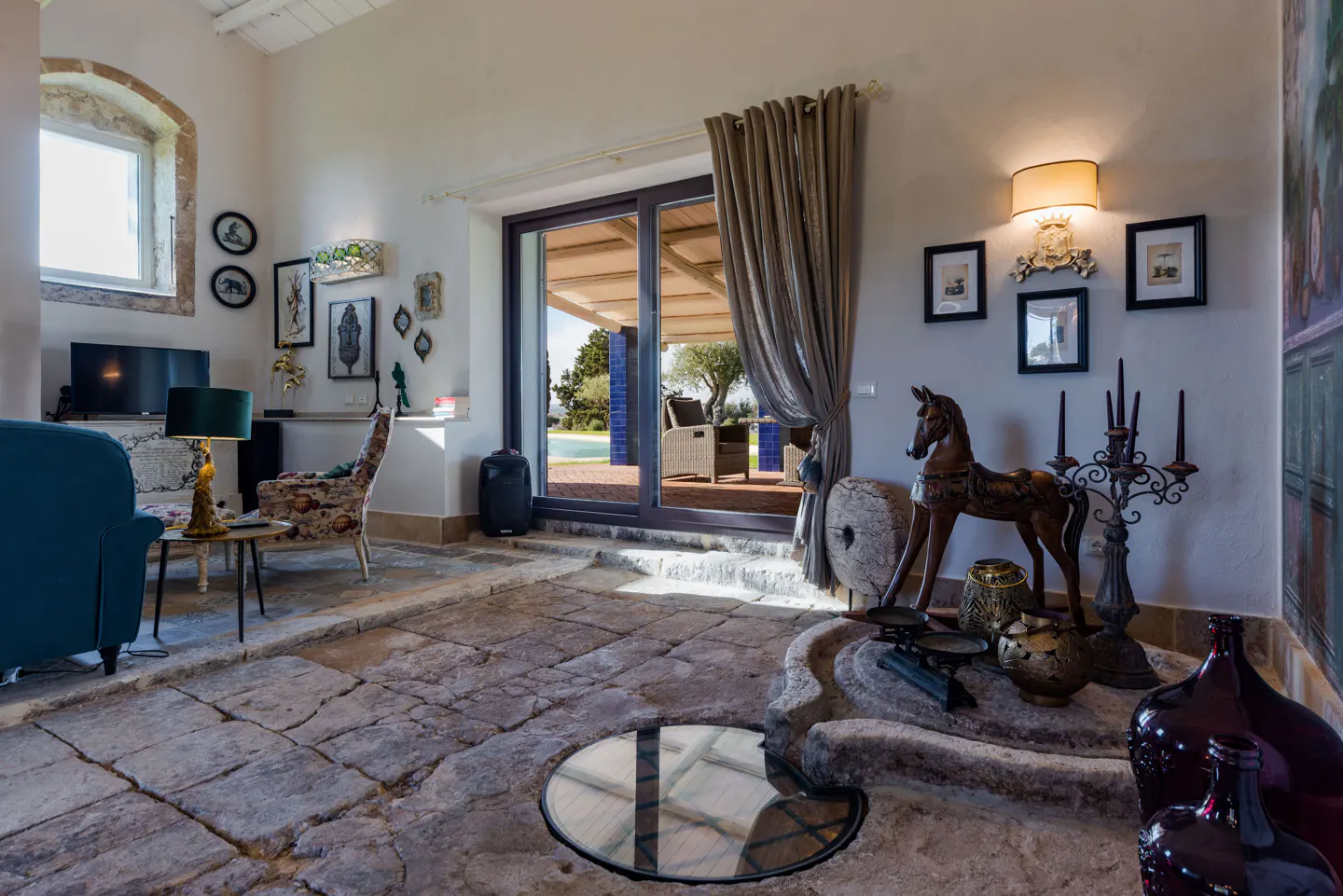 Living room with stone floors, blue sofa, and decor. Sliding glass doors lead to a patio with a pool. A wooden horse statue is near the doors.