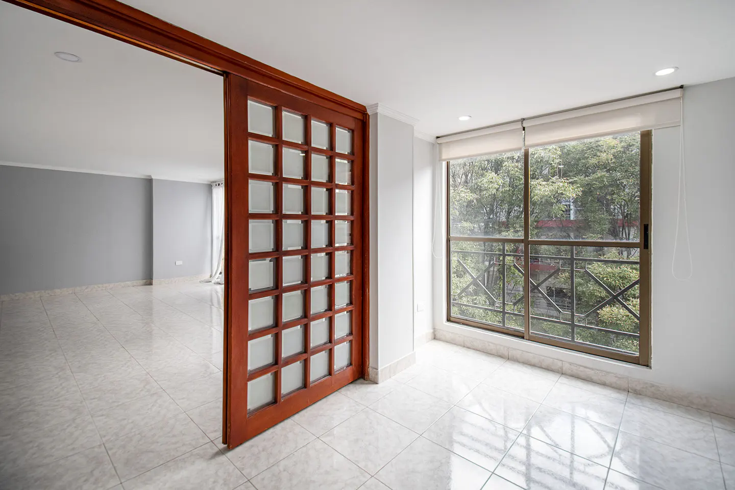 A bright room with a wooden sliding door, white tile floor, and a large window with a view of green trees.