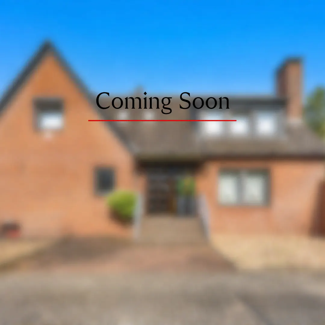 Blurred image of a two-story brick house with a "Coming Soon" banner overlaid on the roofline. Blue sky is visible.