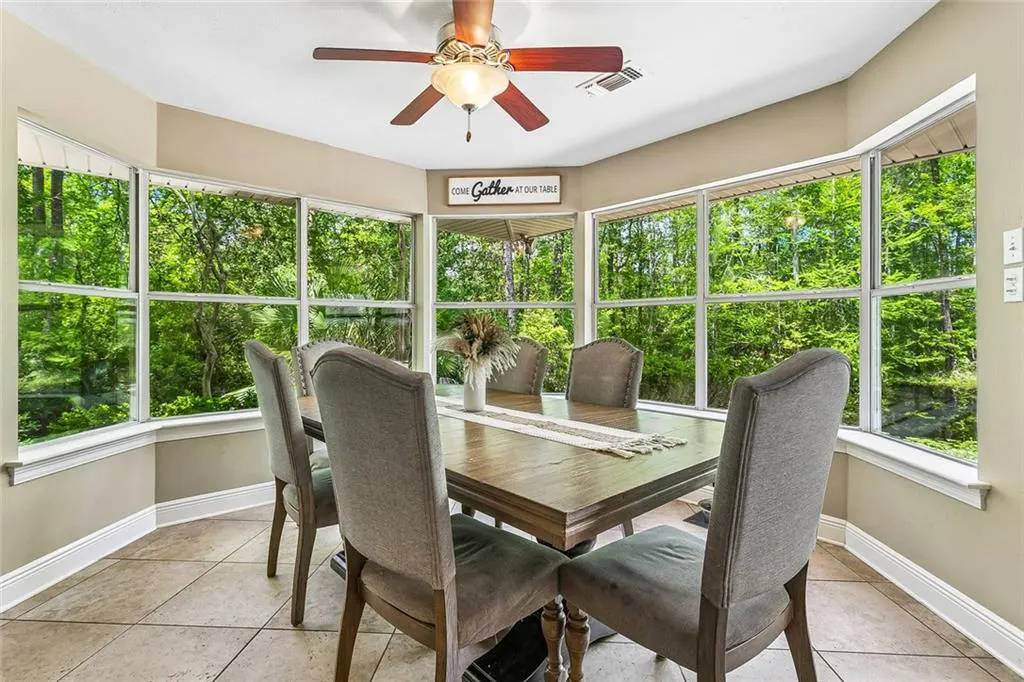 Dining room with a wood table, six gray chairs, and a large bay window overlooking a green forest. A ceiling fan hangs above.