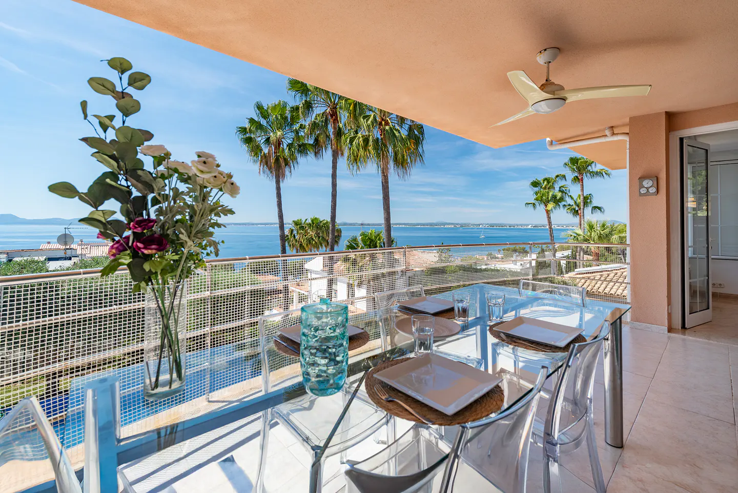 Outdoor dining area with a glass table, clear chairs, and ocean view. Palm trees and blue sky in the background.
