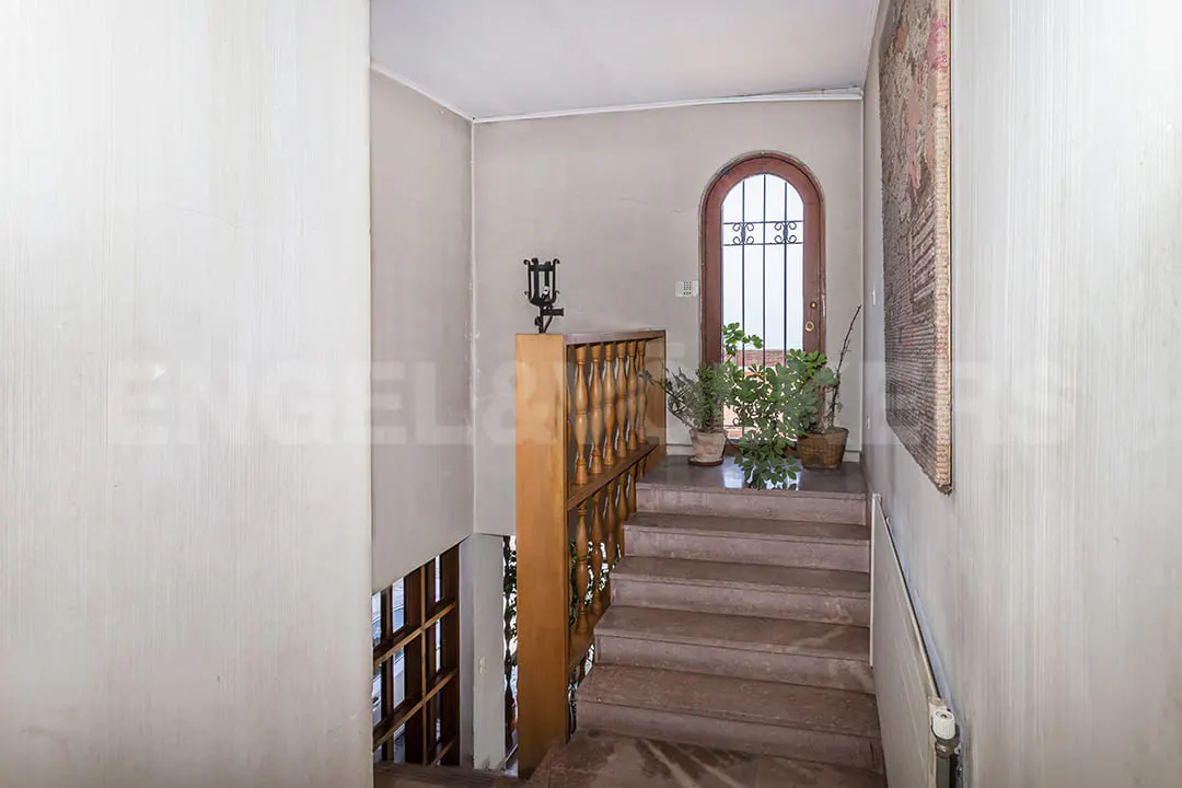 Stairway with wooden railing leads to an arched door with iron bars. Potted plants sit on the landing. A tapestry hangs on the wall.