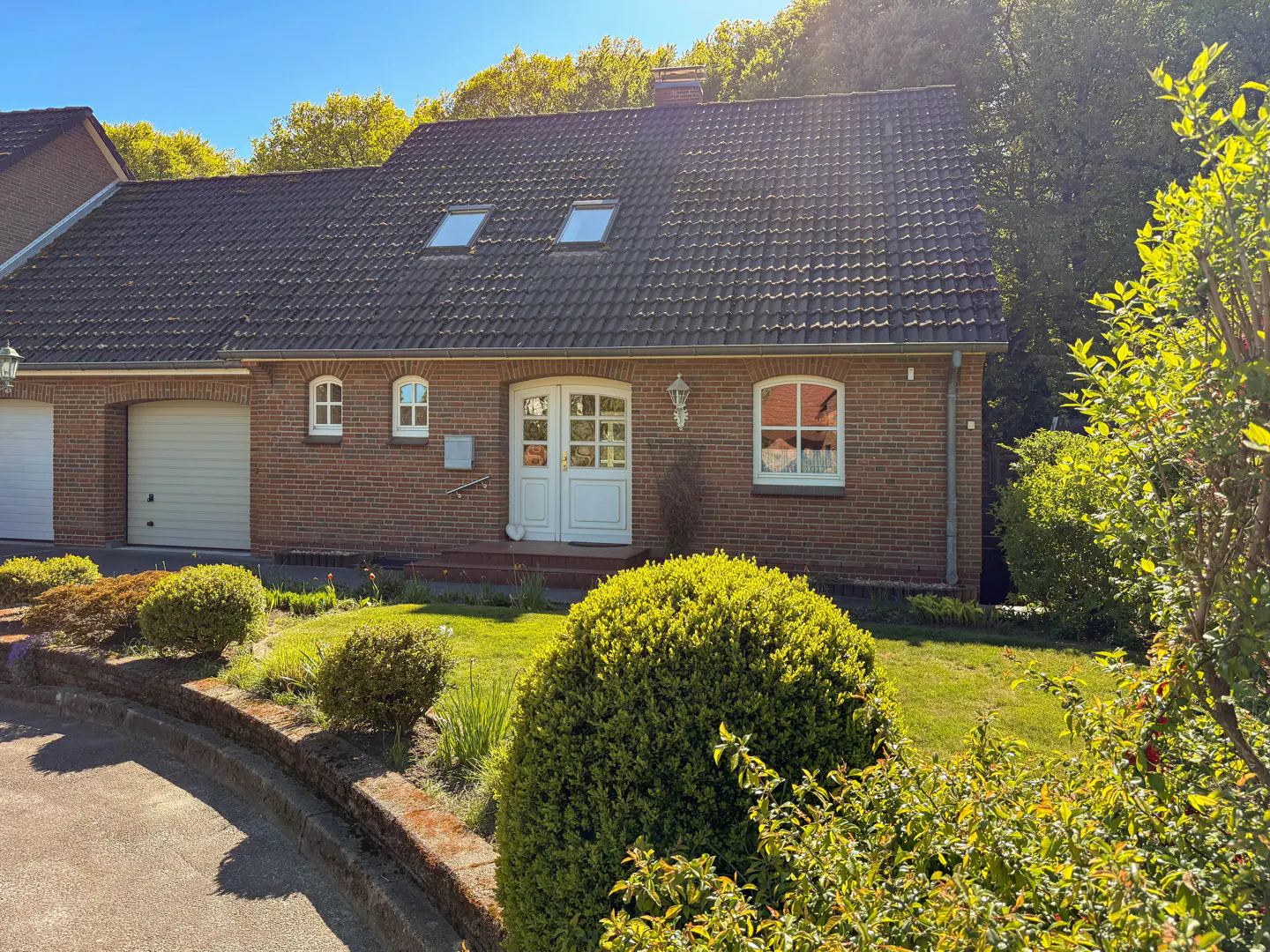 A single-story brick house with a gray tiled roof, white doors, and green bushes in the front yard.