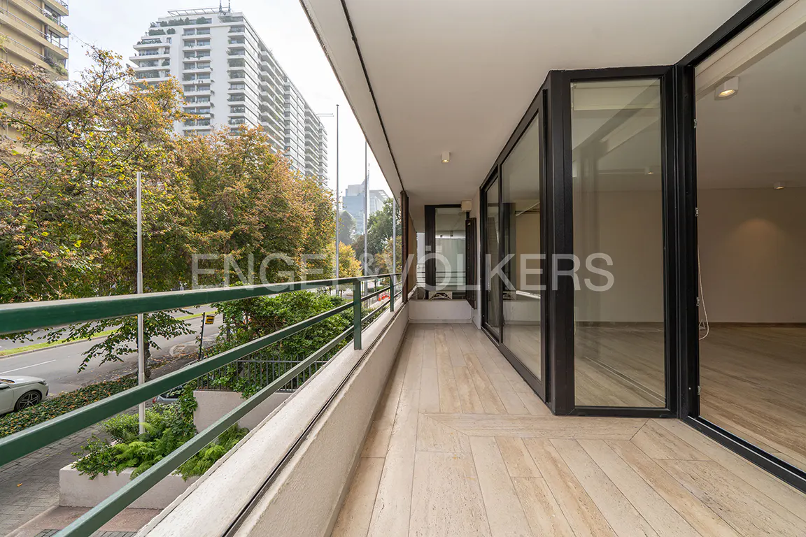 A long balcony with a green railing overlooks a city street with trees and tall buildings. Sliding glass doors lead into the interior.