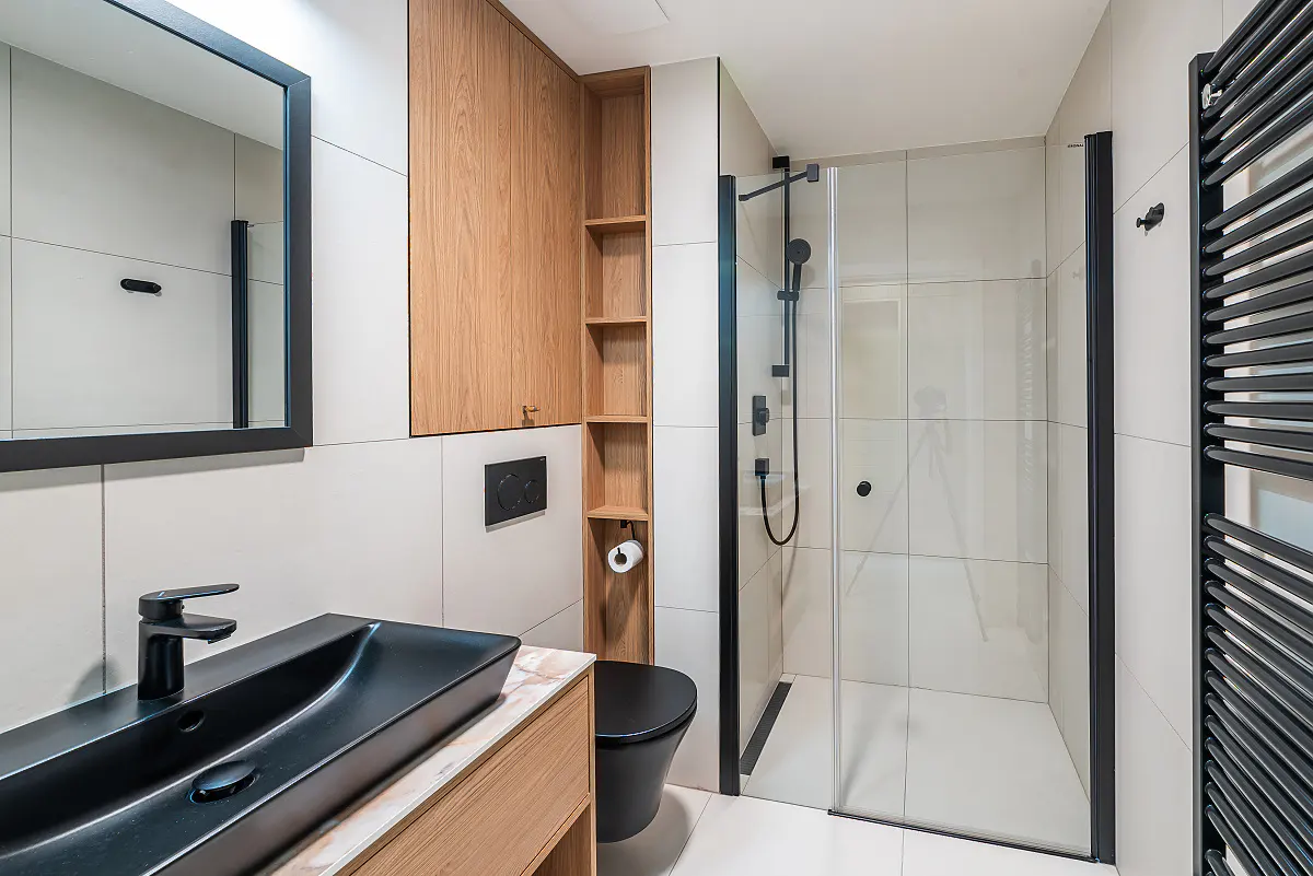Modern bathroom with black fixtures, light wood cabinets, and white tile. A glass shower stall is visible.