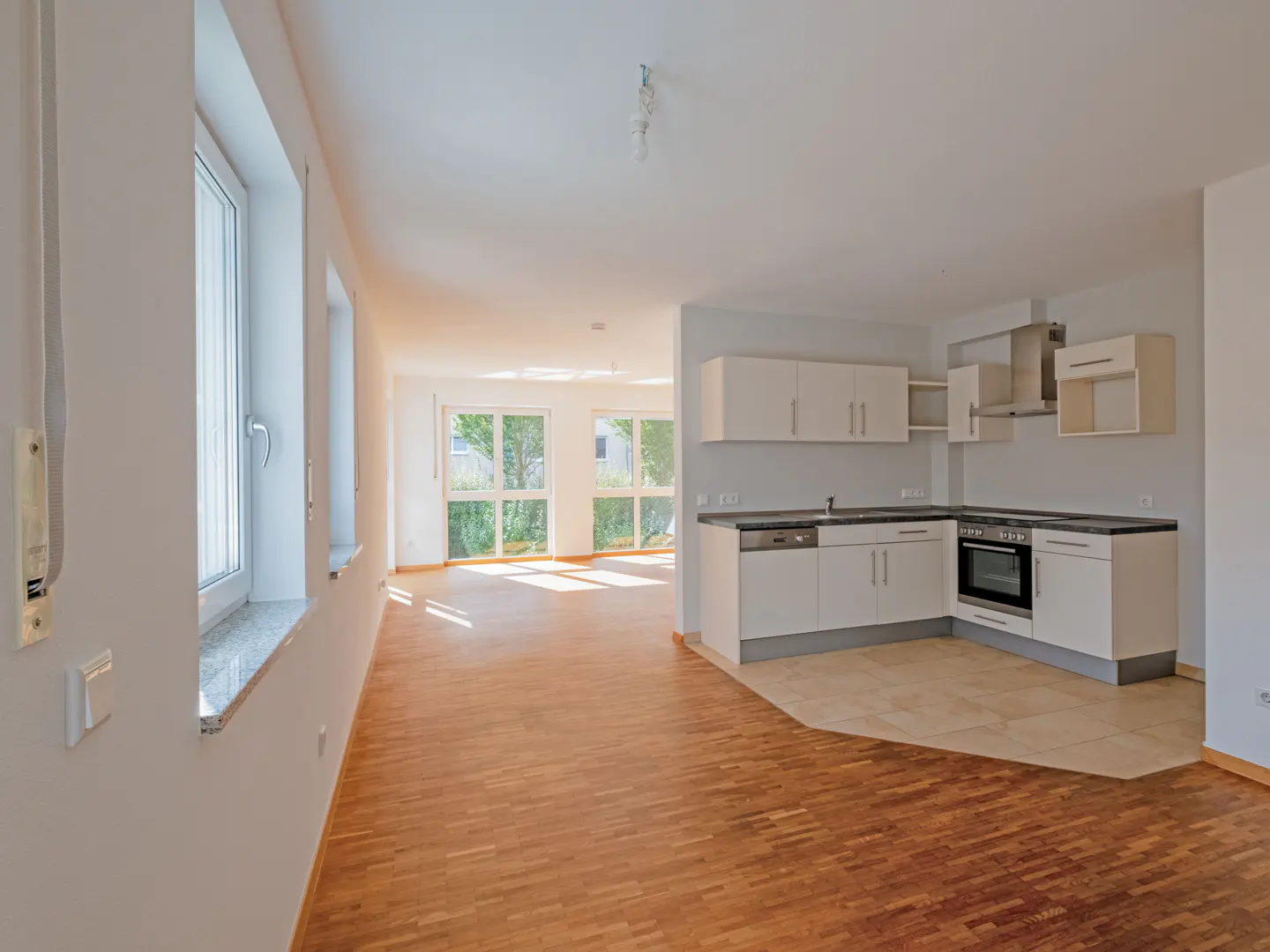 Bright, open-concept apartment with wood floors, white walls, and a modern kitchen with white cabinets. Large windows let in natural light.