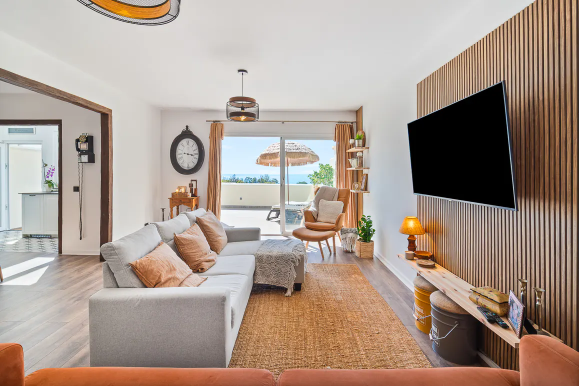 Bright living room with a gray sofa, brown rug, and a large TV on a wood-paneled wall. Balcony with ocean view in the background.