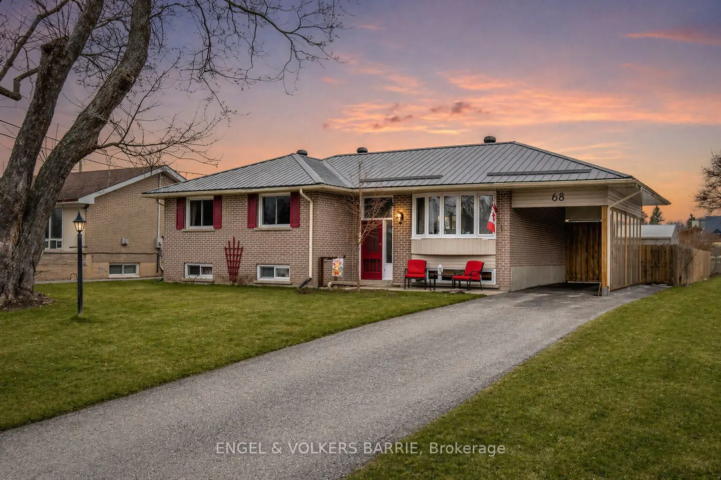 A one-story brick house with a gray roof, red door and shutters, and a long driveway. Two red chairs sit on the porch.