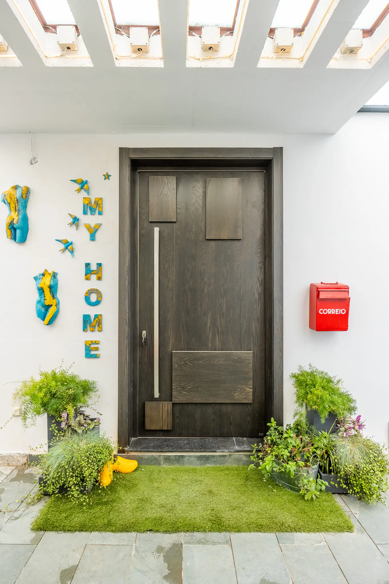 Front entrance with a dark wood door, a red mailbox, and "MY HOME" art on a white wall. Green plants and a green rug add a touch of nature.