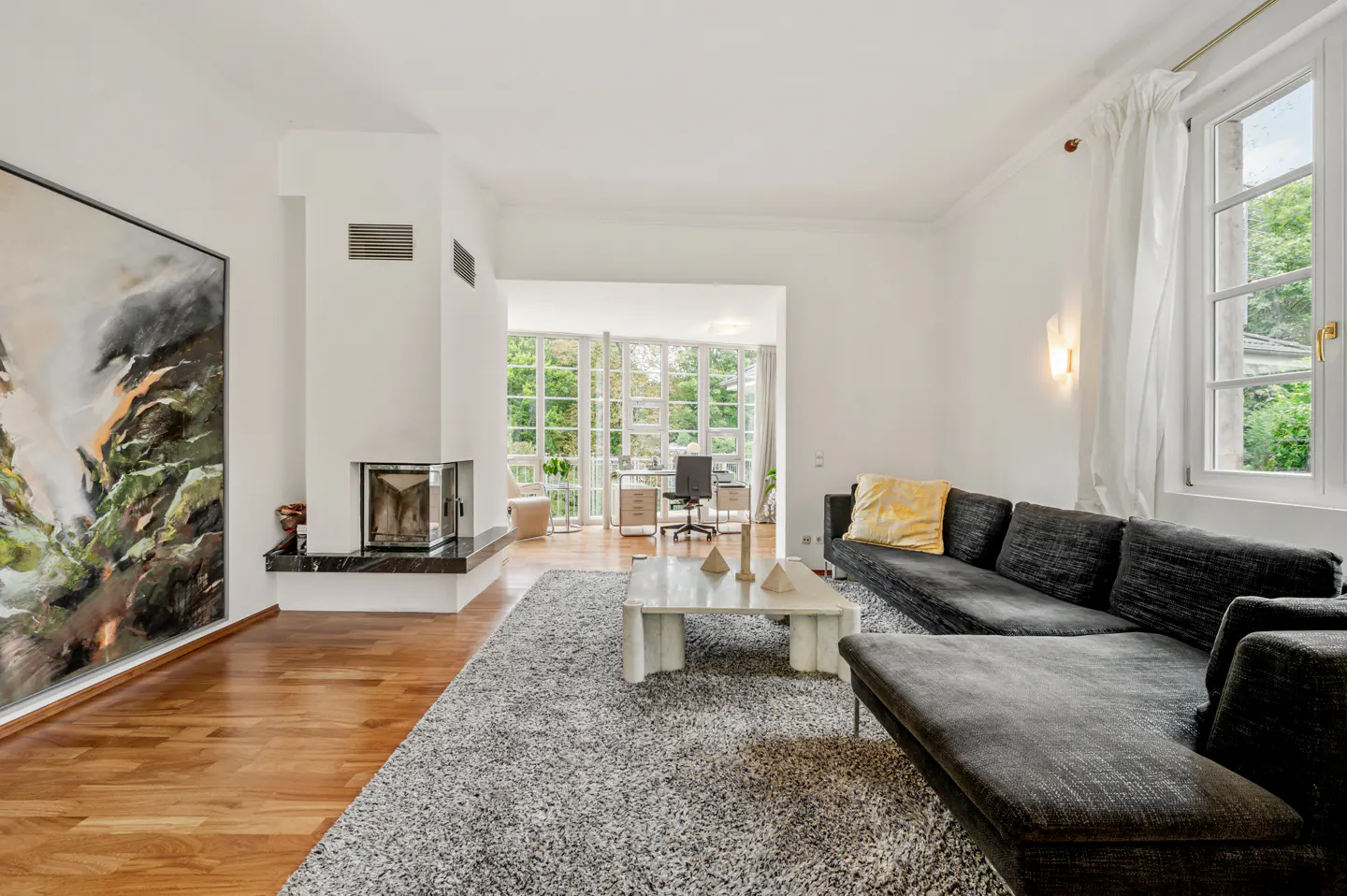 Bright living room with hardwood floors, a gray rug, a black sectional sofa, and a fireplace. A large painting hangs on the wall.