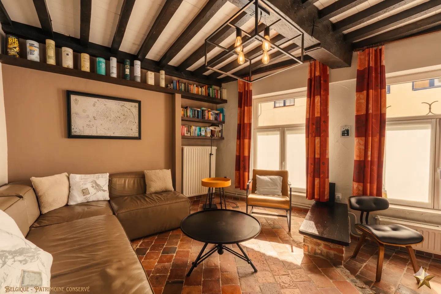Living room with brown leather sofa, black coffee table, and orange curtains. Exposed ceiling beams and brick floor add rustic charm.