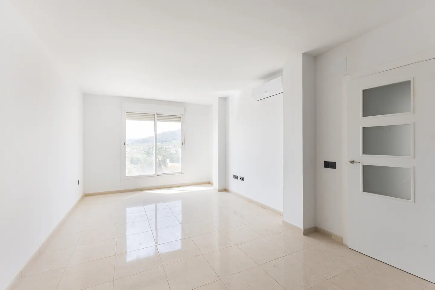 Bright, empty room with white walls, beige tile floor, window with blinds, and a white door with three glass panels.