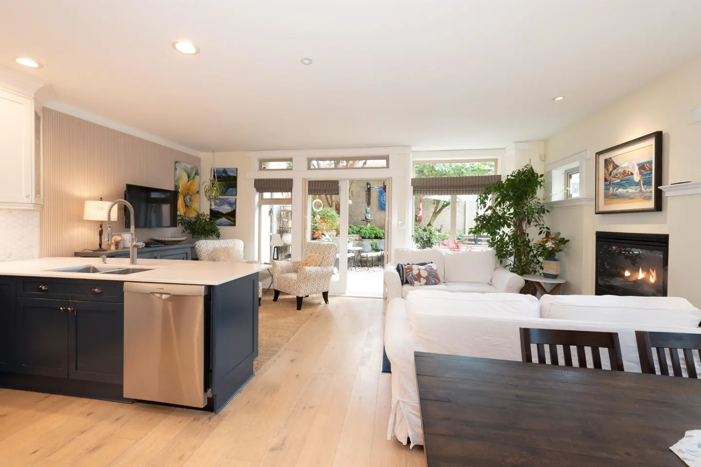 Open-concept kitchen and living area with dark blue cabinets, white sofa, fireplace, and patio doors leading to a garden.
