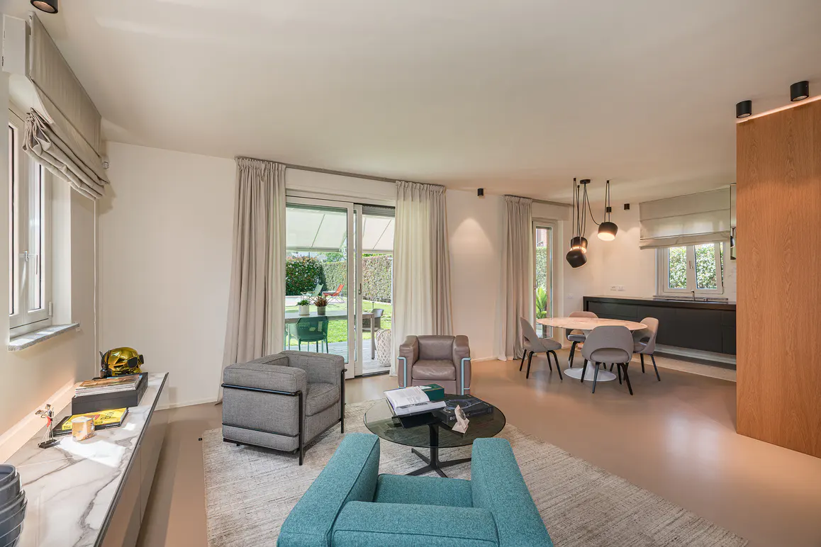 Bright living room with blue and gray armchairs, a round glass table, and a dining area with a round table and gray chairs. Sliding glass doors lead to a green lawn.