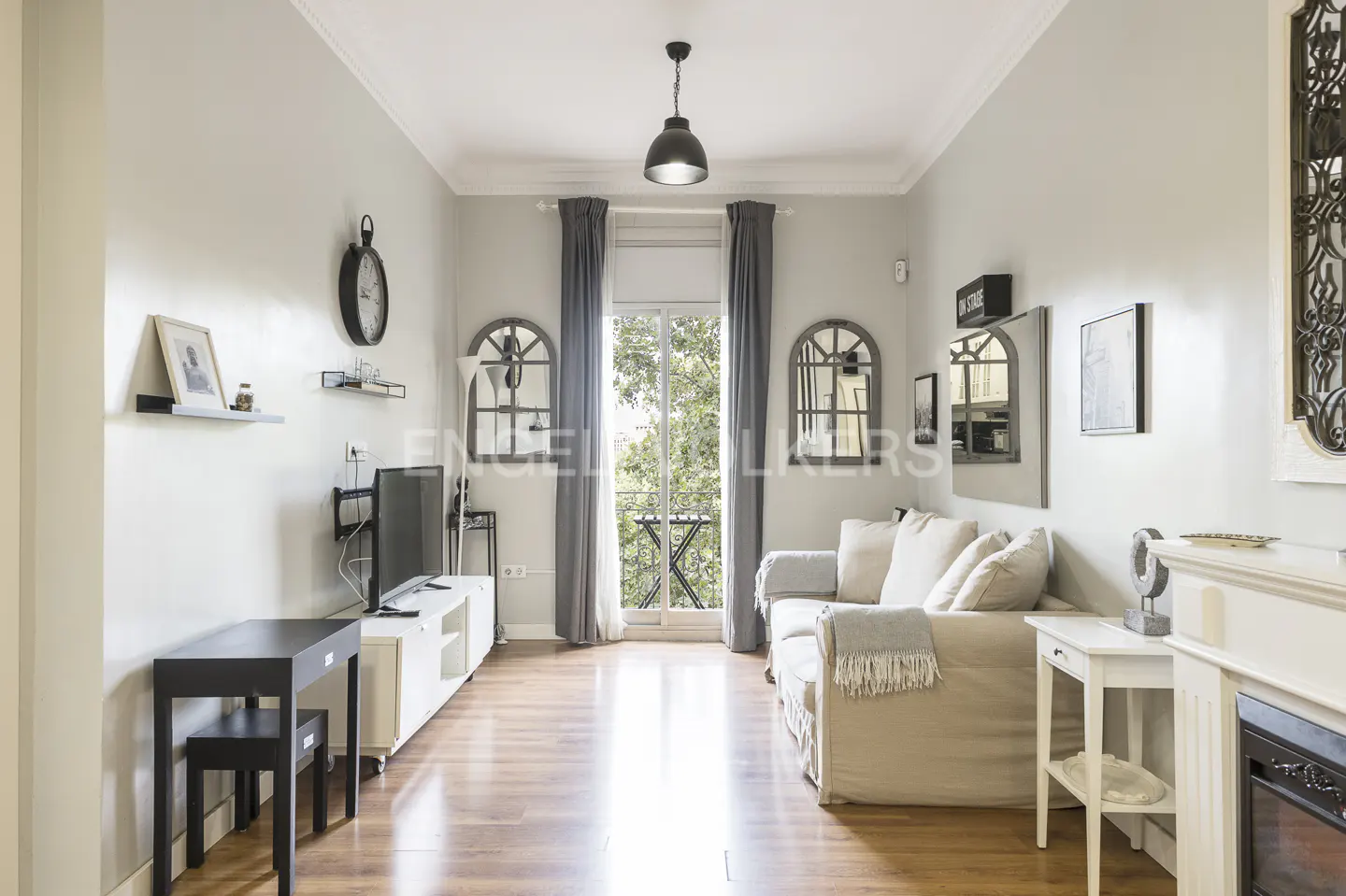 Living room with hardwood floors, light gray walls, and a beige sofa. A black desk and TV stand are on the left. Balcony doors with gray curtains are centered.