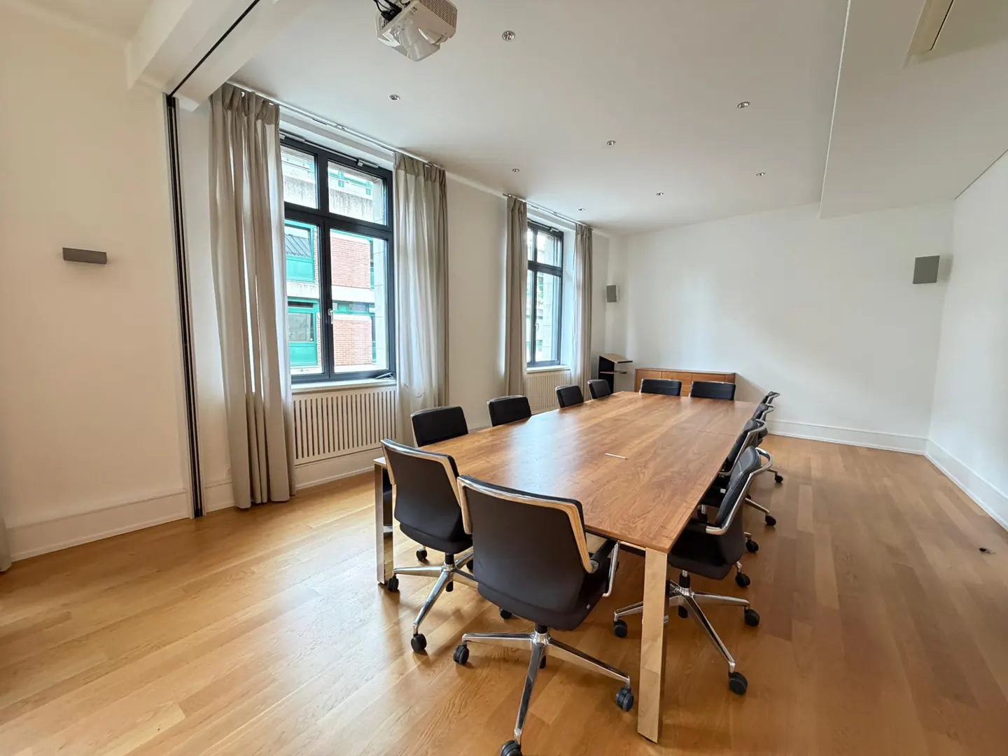 Bright conference room with a long wooden table, black chairs, and large windows with light curtains. A projector is mounted on the ceiling.