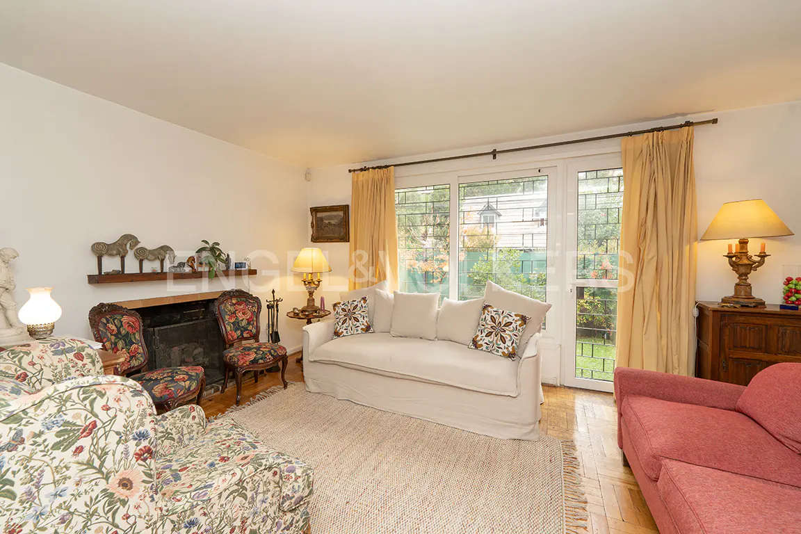 Living room with floral armchairs, a beige sofa, and a fireplace. A window with yellow curtains overlooks a green garden.