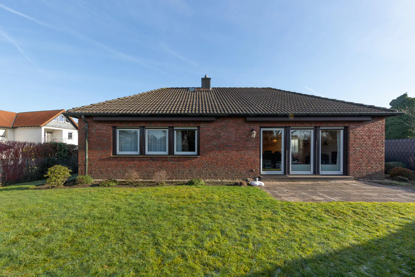 Back view of a one-story red brick house with a brown tiled roof, a green lawn, and a stone patio.