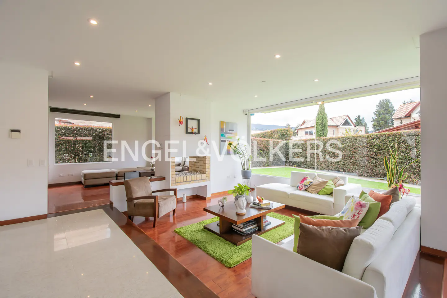 Bright living room with white sofas, colorful pillows, and a wooden coffee table on a green rug. Large windows overlook a green lawn.