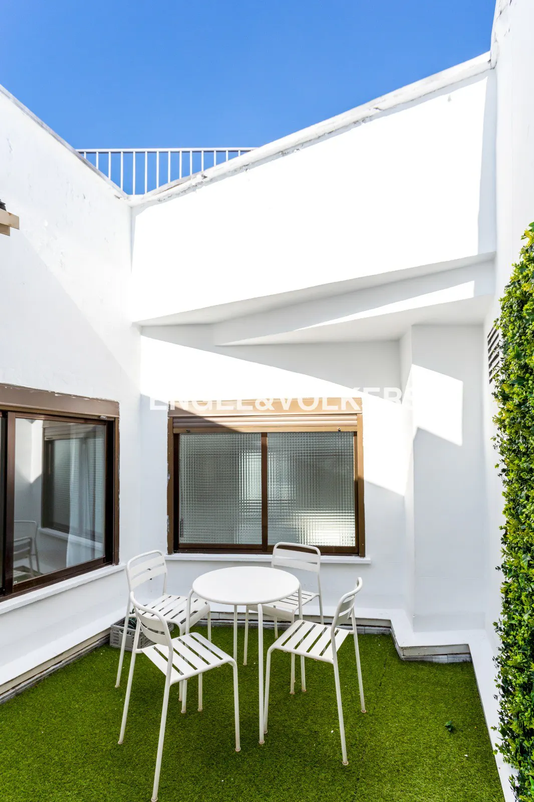 White patio set with a round table and four chairs on a green artificial grass terrace against white walls under blue sky.