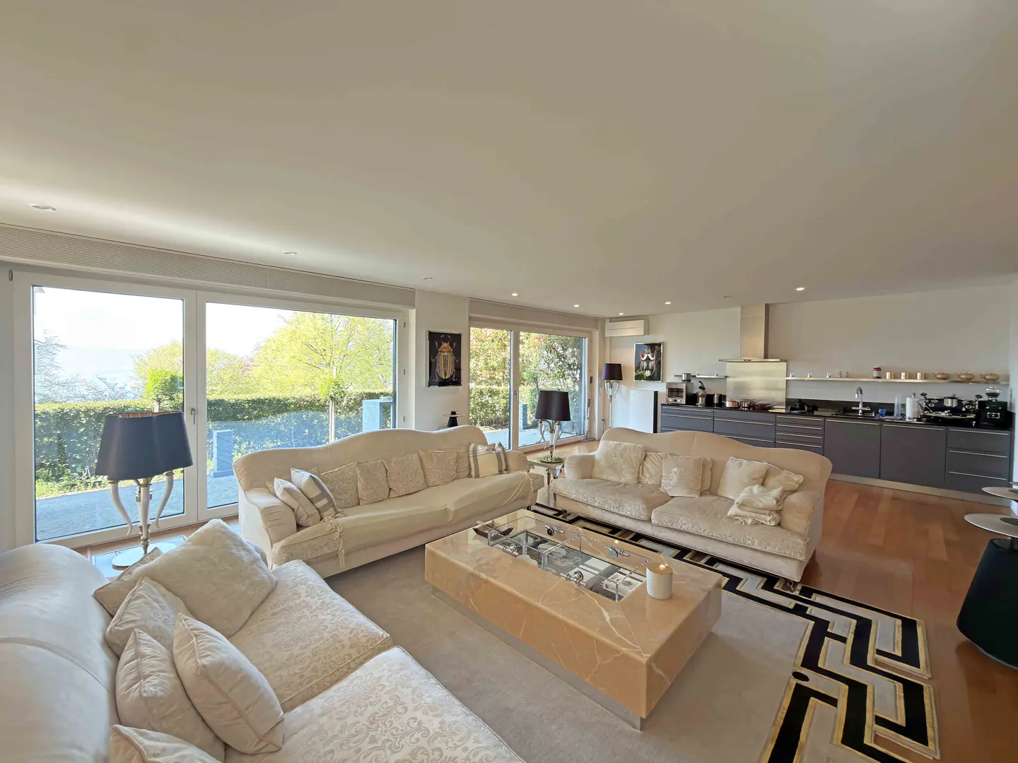 Bright living room with cream sofas, marble coffee table, and a view of greenery through large windows. Kitchen in the background.