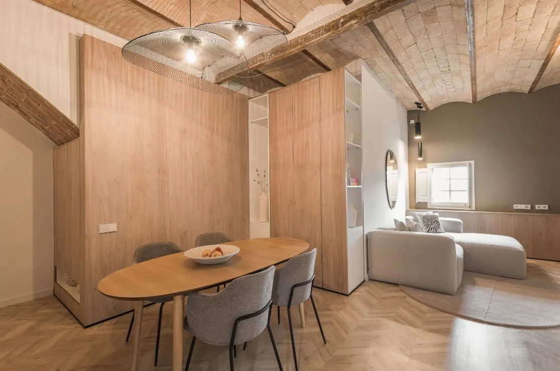 A modern apartment interior with a dining table, gray chairs, and a white sofa. The room features a herringbone floor and a vaulted brick ceiling.