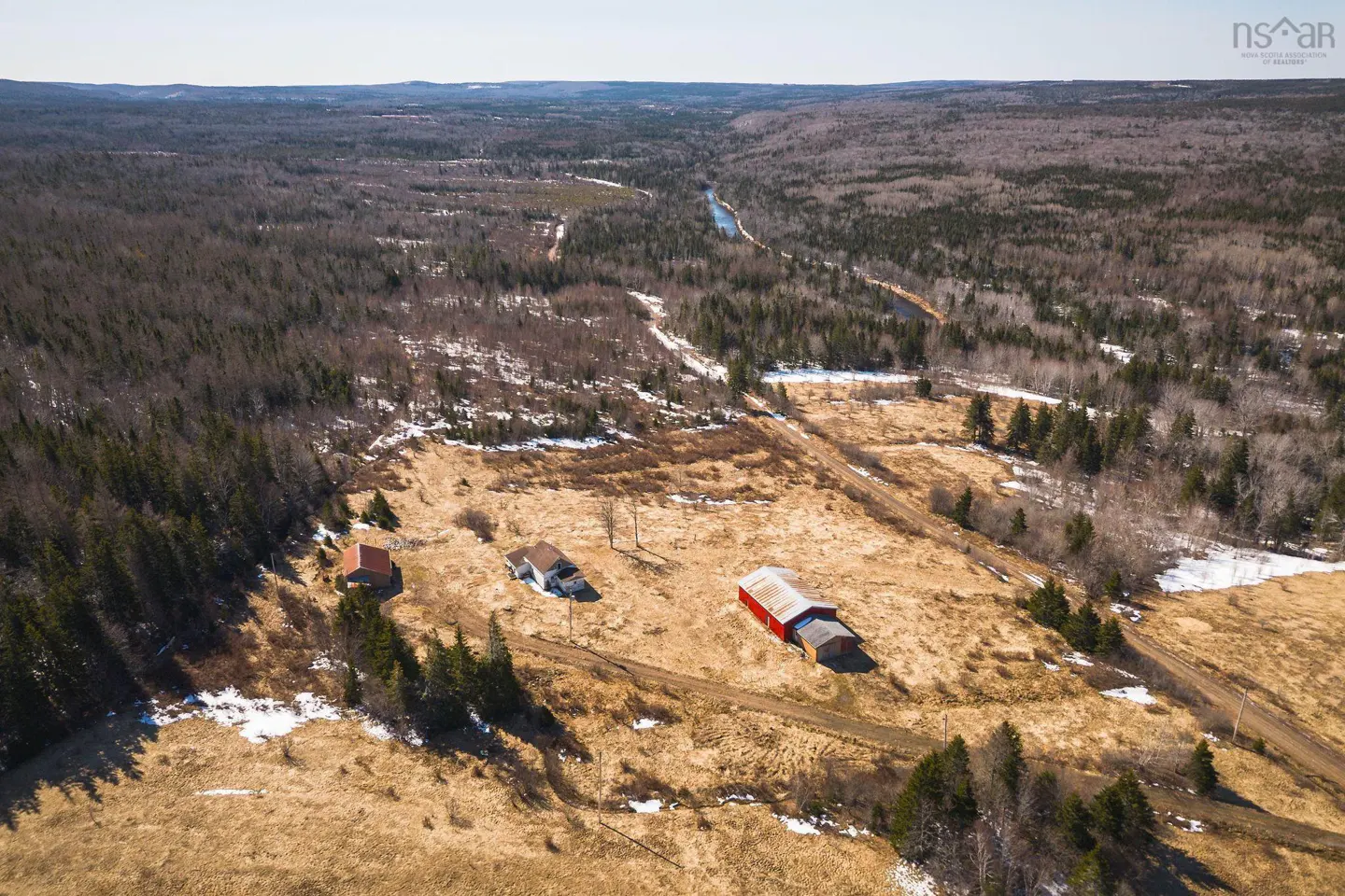 Aerial view of a rural property with a red barn, house, and outbuilding surrounded by fields and forest.