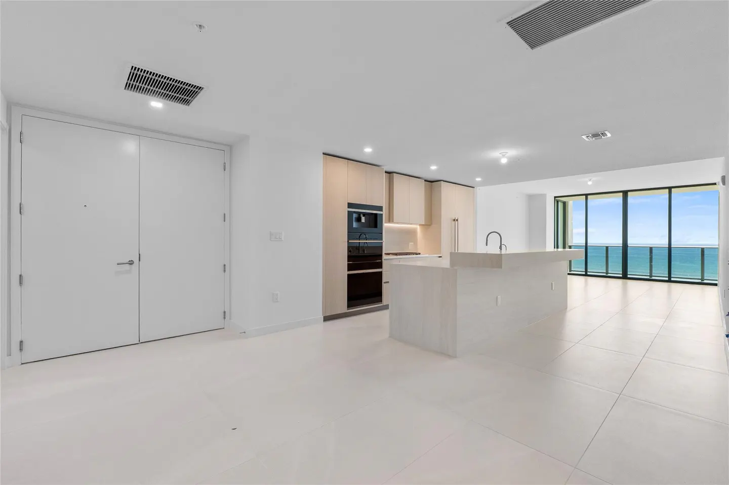 Bright, modern condo interior with white tile floors, kitchen island, and ocean view through large windows.