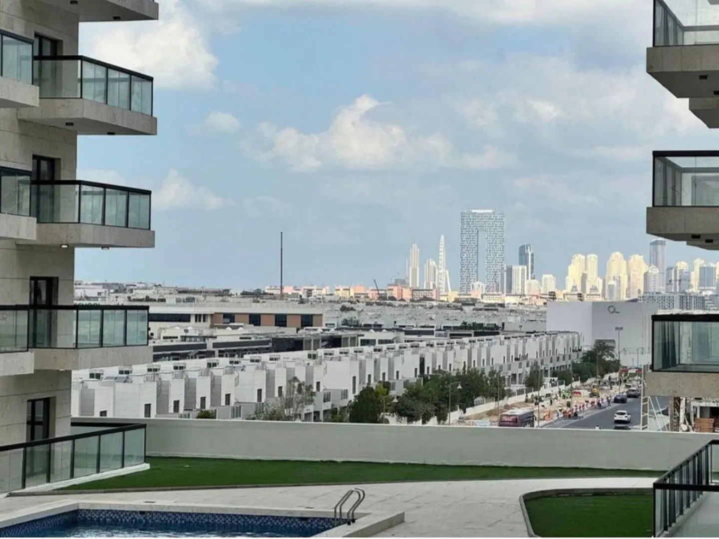 View from a balcony with a pool, overlooking white townhouses and the Dubai skyline under a cloudy sky.