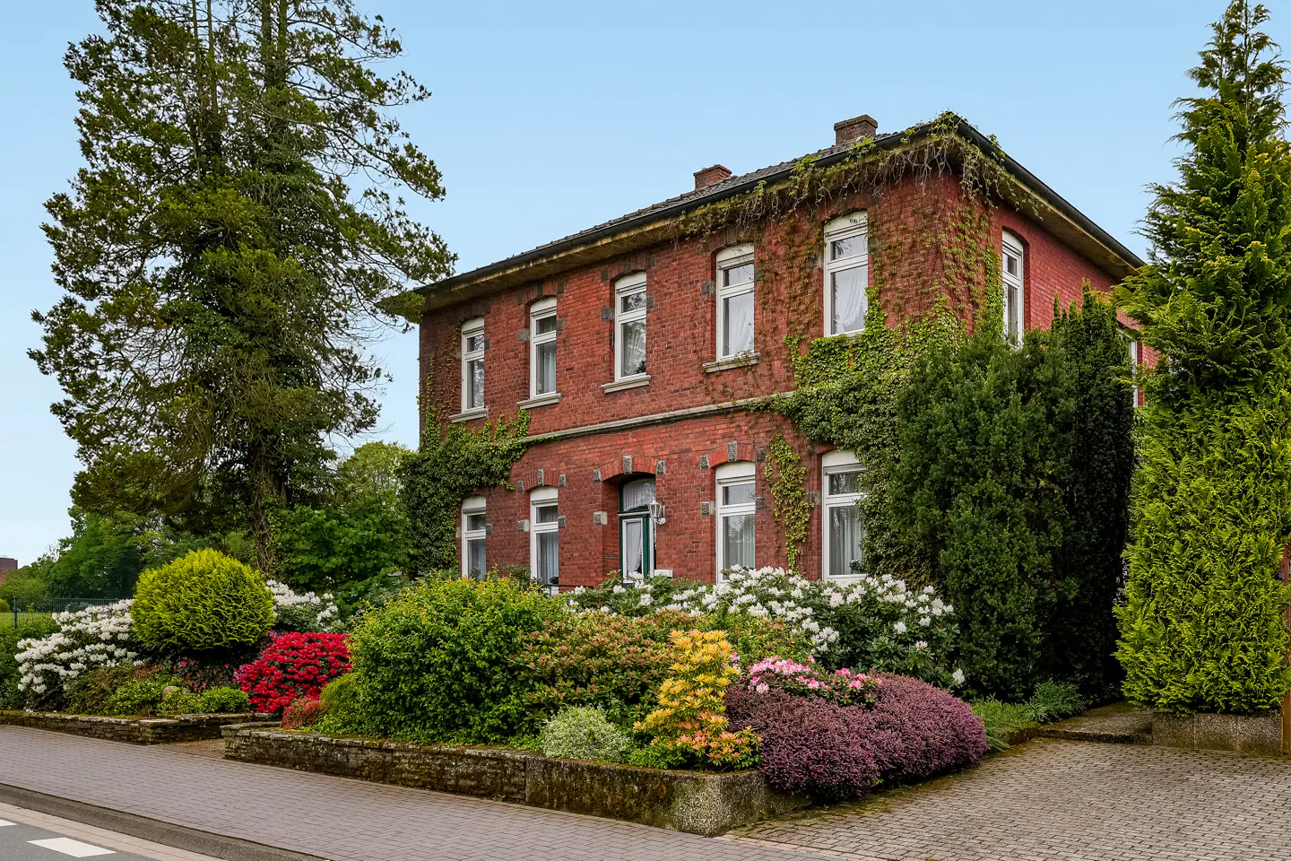 Two-story red brick house with white-framed windows, partially covered in ivy. Colorful flower garden in front.