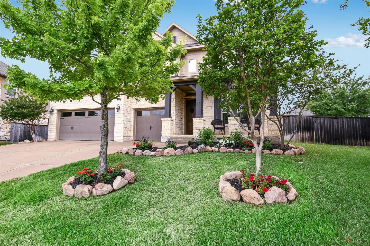 Two-story stone house with a green lawn, trees, and flower beds. The house has a two-car garage and a covered front porch with chairs.
