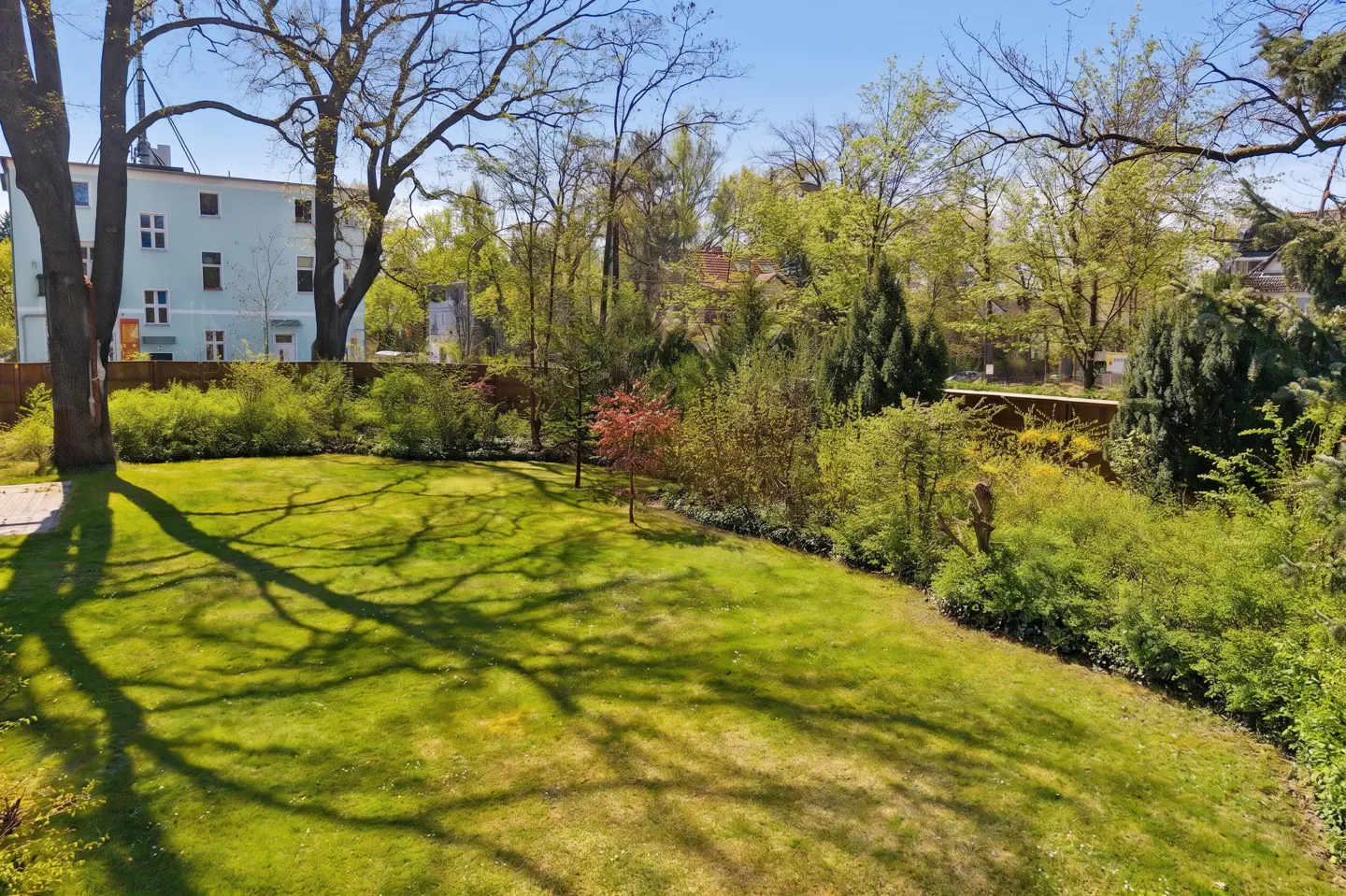 A lush green lawn with tree shadows, surrounded by trees and shrubs. A light blue building is visible in the background.