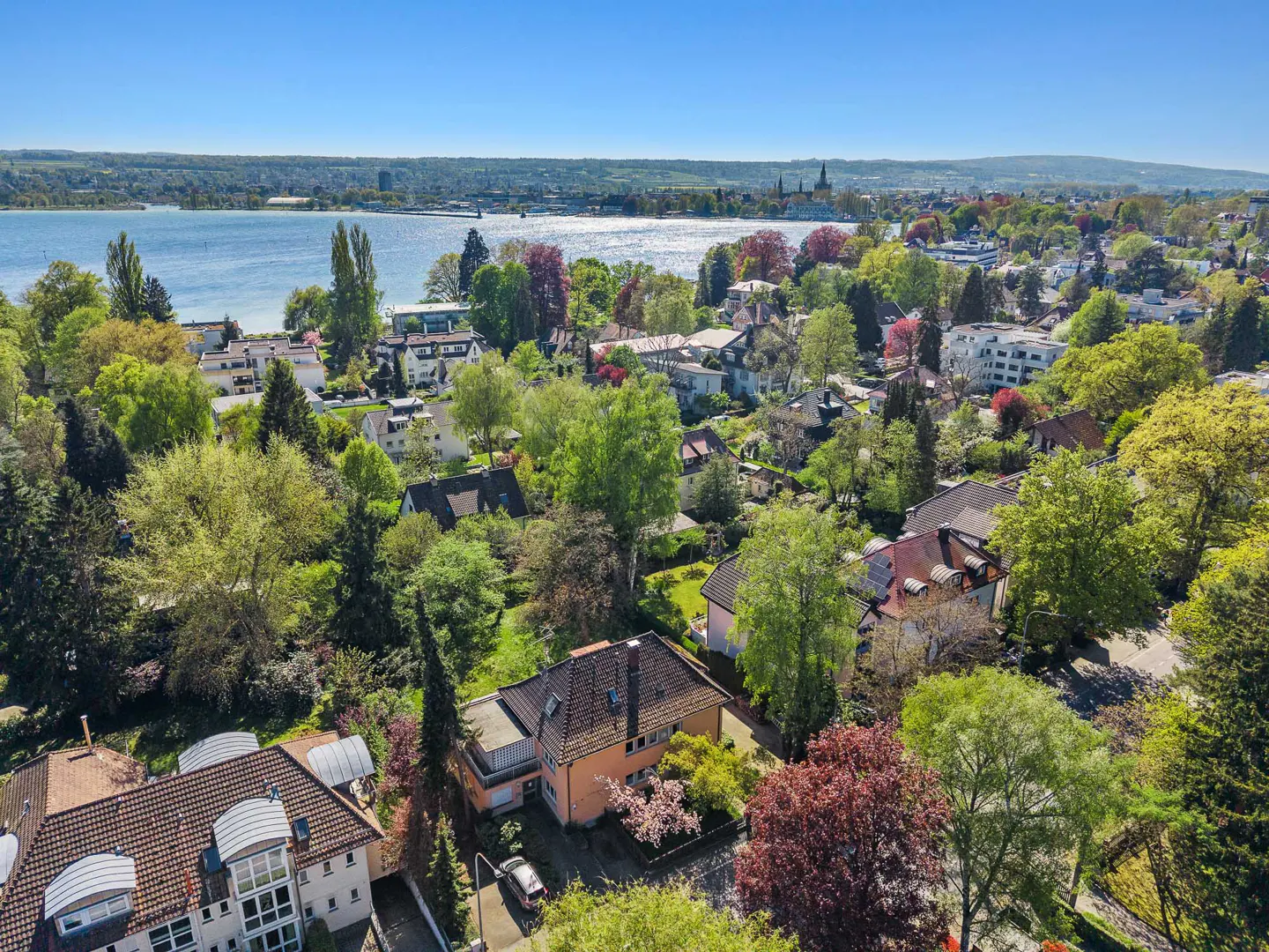 Aerial view of a residential area with houses, trees, and a body of water in the background under a clear blue sky.