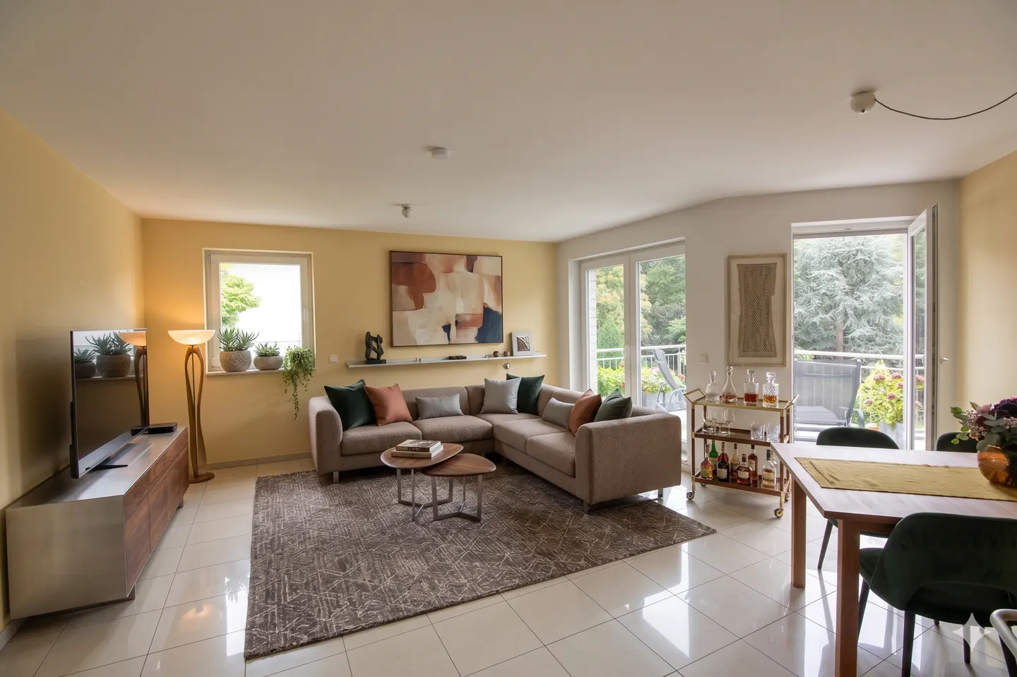 A bright living room with a beige sectional sofa, a wooden dining table with green chairs, and a bar cart.