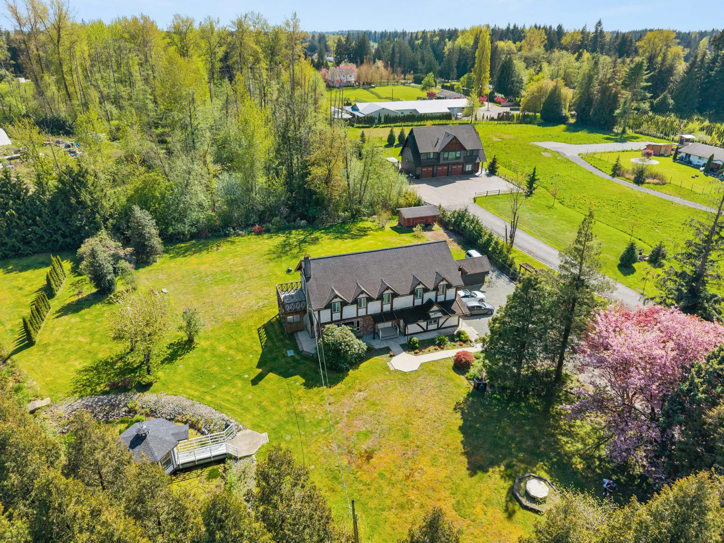 Aerial view of a large, two-story white and brown house with a black roof, surrounded by green lawns, trees, and a pond.