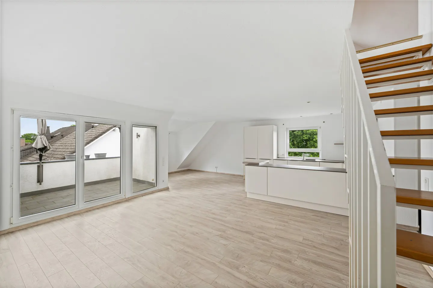 Bright, open-plan apartment with light wood floors, white walls, kitchen, balcony access, and wooden stairs with white railings.