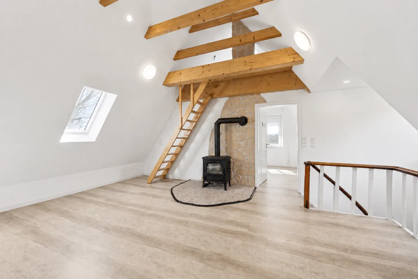 Attic room with white walls, wood beams, and a black wood-burning stove. A wooden ladder leads to the beams. A skylight is on the left.