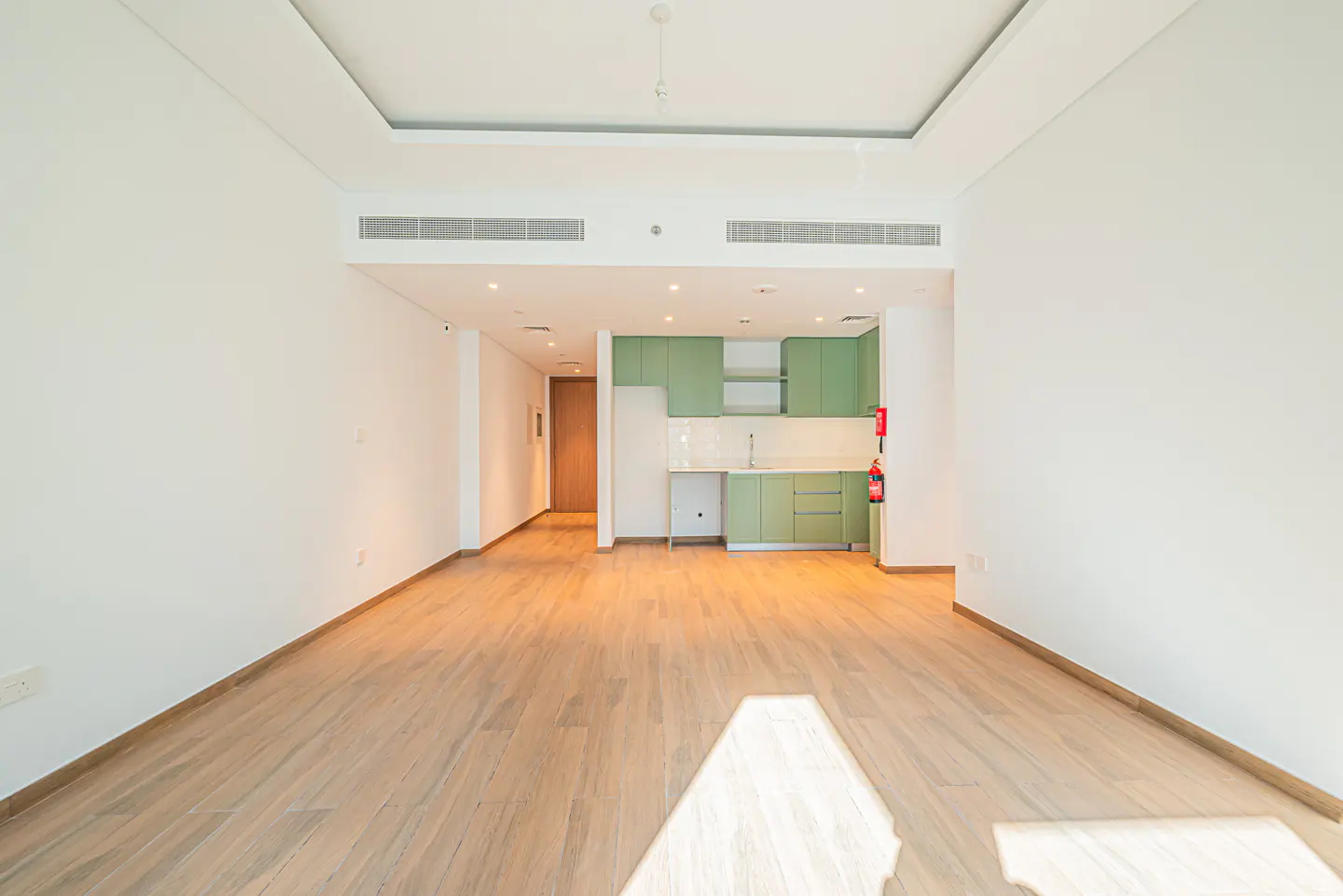 Bright, empty apartment with light wood floors, white walls, and a green kitchen in the background.